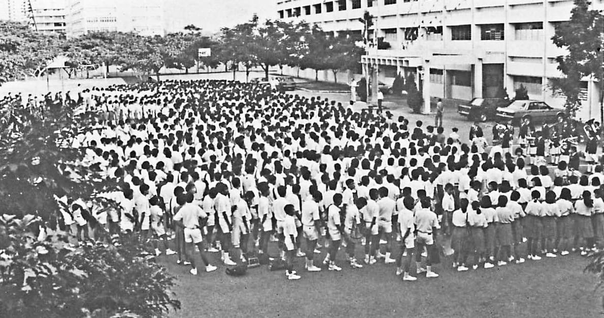 Large crowd of students in white uniforms gathered on a school field near a multi-story building, black and white photo.
